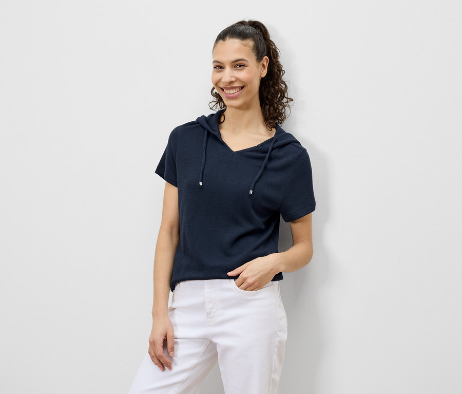Frau mit lockigem Haar lächelt in marineblauem Kapuzen-T-Shirt und weißen Hosen.