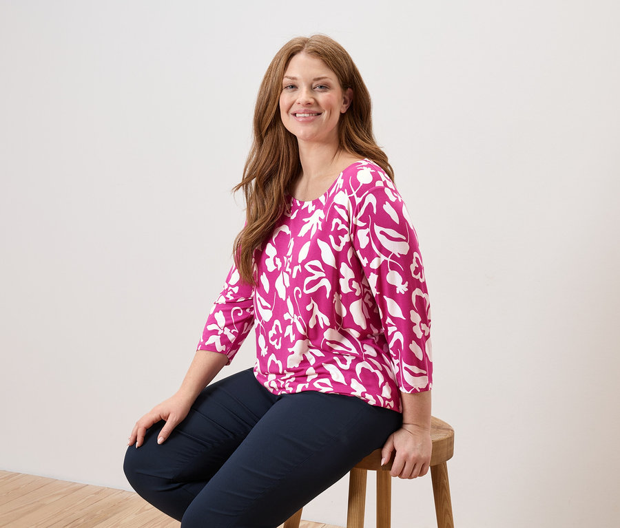 Eine Frau mit langen, roten Haaren sitzt auf einem Holzstuhl und trägt eine pinkfarbene Bluse mit weißen Blättern und marineblaue Hosen.