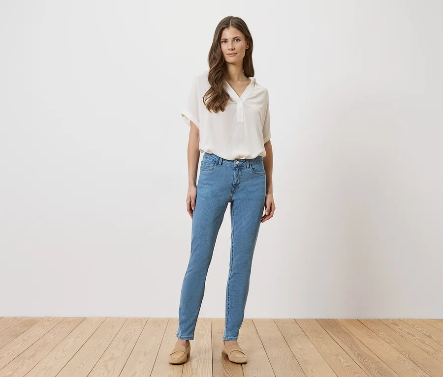Frau in hellblauen Jeans, beiger Bluse und Schuhen steht vor einer weißen Wand auf einem Holzboden.