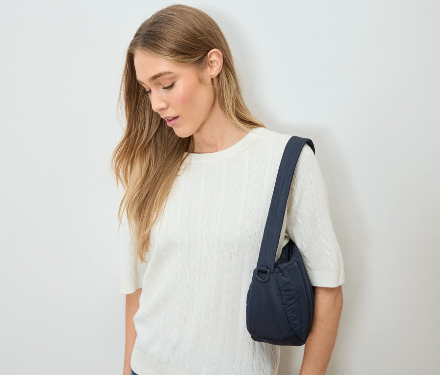 Frau mit blonden Haaren und weißem Pullover trägt eine marineblaue Handtasche über der Schulter.