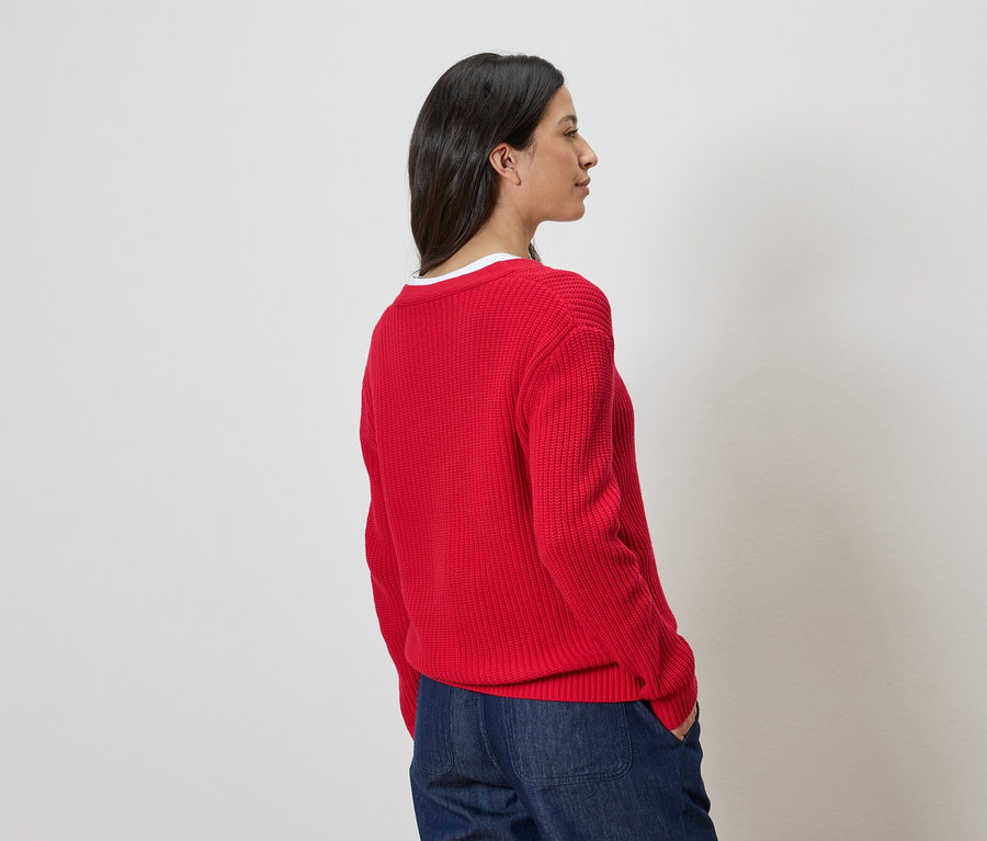 Frau von der Seite, trägt einen roten Strickpullover und blaue Jeans.