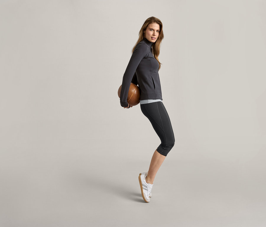 Frau hält einen braunen Ball hinter ihrem Rücken, trägt eine graue Jacke und 2 Tops, schwarz/grau.