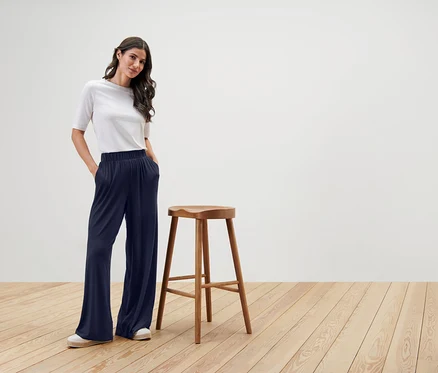 Frau steht neben einem Holzstuhl, trägt ein weißes T-Shirt und eine weite, dunkelblaue Hose.