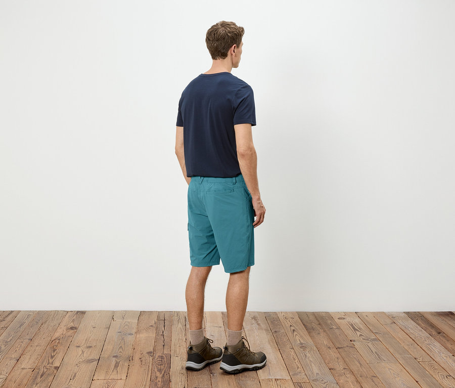 Mann von hinten, mit blauem T-Shirt, türkisfarbenen Shorts und Wanderschuhen.