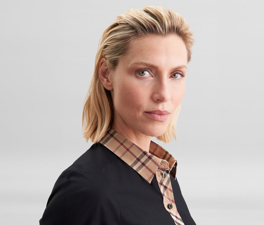 Nahaufnahme einer Frau mit blondem Haar, die eine schwarze Seidensticker Popeline-Bluse mit beigem Karodetail am Kragen trägt.