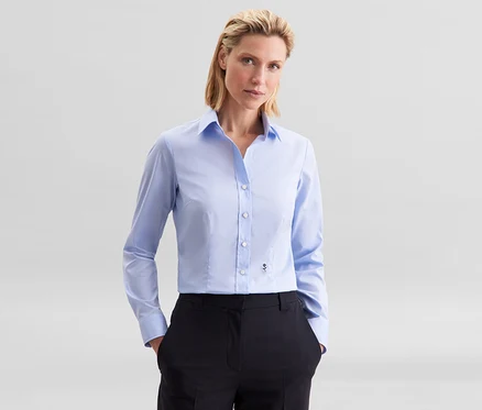 Eine Frau trägt eine hellblaue Seidensticker Slim Fit non iron Bluse und schwarze Hose, die Hände in den Taschen.