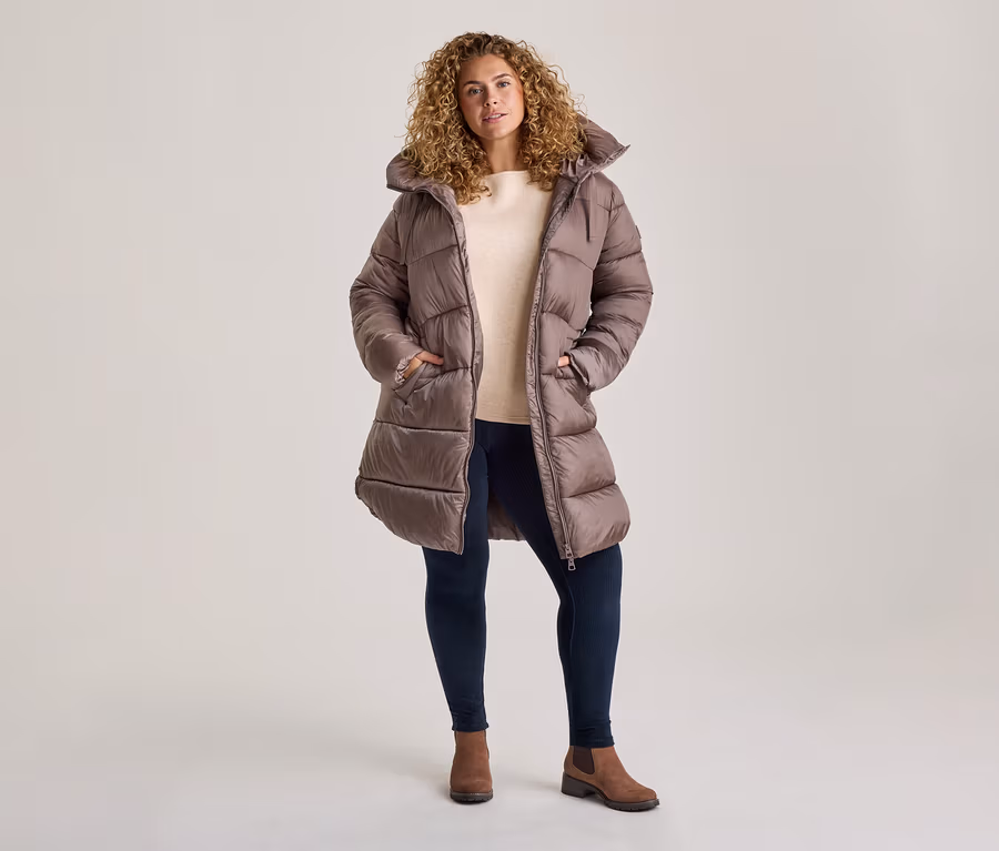 Frau mit lockigem Haar trägt einen langen braunen Steppmantel, ein helles Oberteil, dunkle Hose und braune Stiefeletten.
