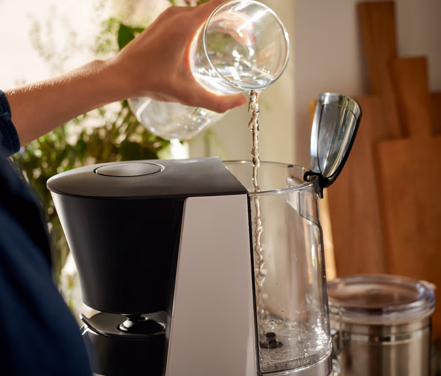 Frau gießt Wasser aus einem durchsichtigen Glas in eine grau-schwarze »Let's Brew« Filterkaffeemaschine.