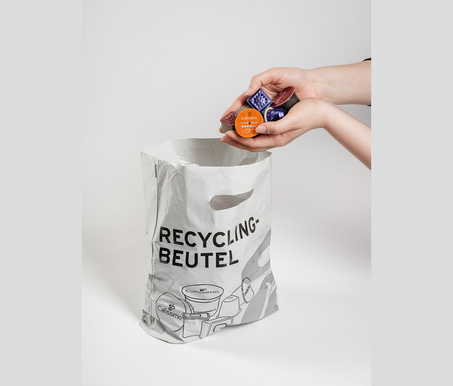 Hände halten bunte Kaffeekapseln, darunter eine mit „Cafissimo Caffe Crema“, über einem weißen Recycling-Beutel mit Aufschrift „RECYCLING-BEUTEL“ und Kapselillustrationen.