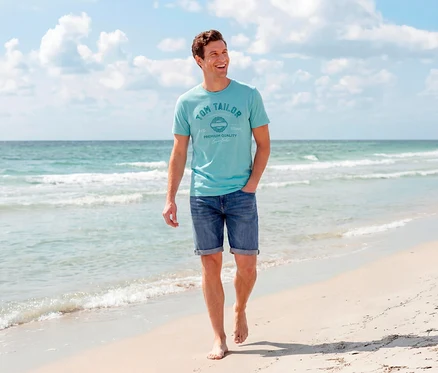 Ein lächelnder Mann geht barfuß am Sandstrand, trägt ein türkises Tom Tailor T-Shirt mit Logo und mitteldunkle Tom Tailor TTJOSH REGULAR SLIM Jeans Shorts, mit Meer und blauem Himmel im Hintergrund.