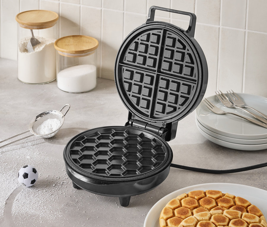 Offenes, schwarzes Waffeleisen auf einer Küchentheke mit fertigen Waffeln auf einem Teller, einem kleinen Fußball und Zutaten in Gläsern im Hintergrund.