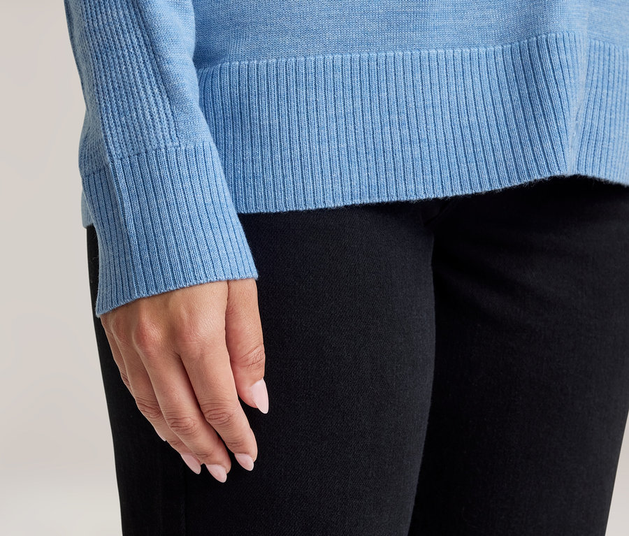 Nahaufnahme einer Person in einem hellblauen Pullover mit gerippten Bündchen und Saum sowie einer schwarzen Hose. Die Hände der Person mit hellrosa Nagellack sind sichtbar.