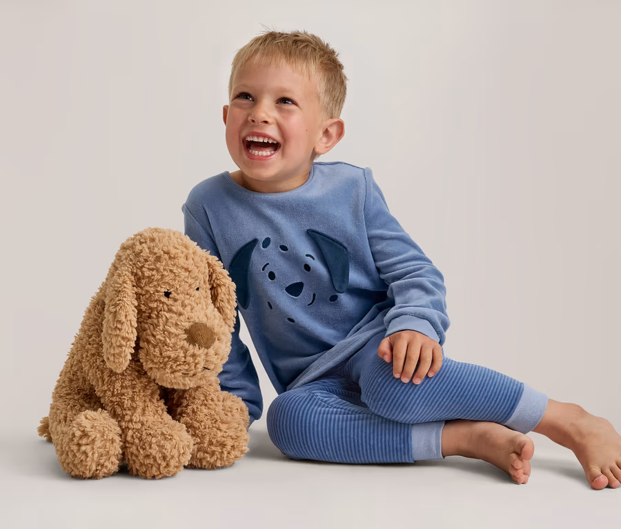 Ein lachender kleiner Junge in blauem Pyjama mit Hunde-Gesichtsmotiv sitzt neben einem flauschigen braunen Plüschhund.