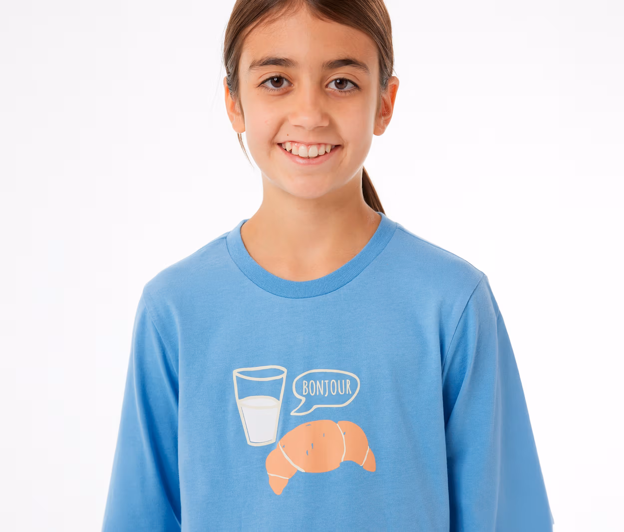 Lächelndes junges Mädchen in einem blauen SCHIESSER Kinder-Pyjama-Oberteil mit einem Aufdruck aus Milchglas, Croissant und einer Sprechblase mit „BONJOUR“.