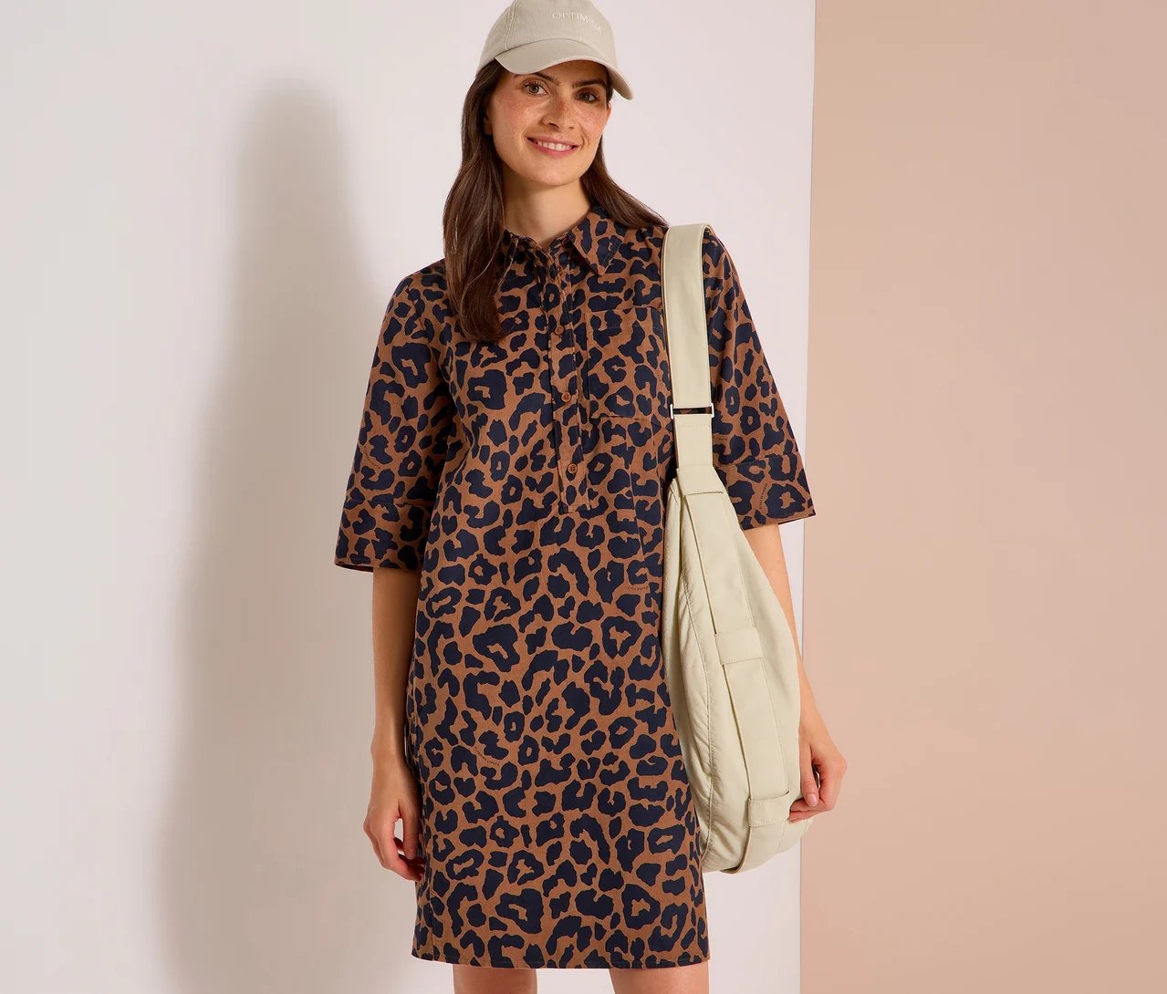 Lächelnde Frau in braun-schwarzem Leopardenkleid, beiger Baseballkappe und beiger Umhängetasche.