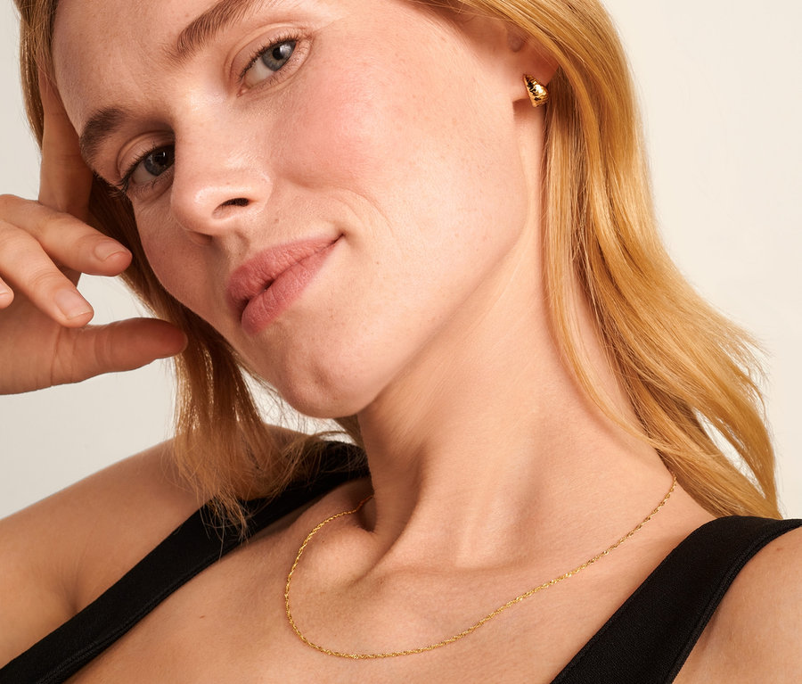 Blonde Frau mit Hand am Kopf trägt eine 585 Gold Kette Singapur und 375 Gold Halbcreolen konisch diamantiert.