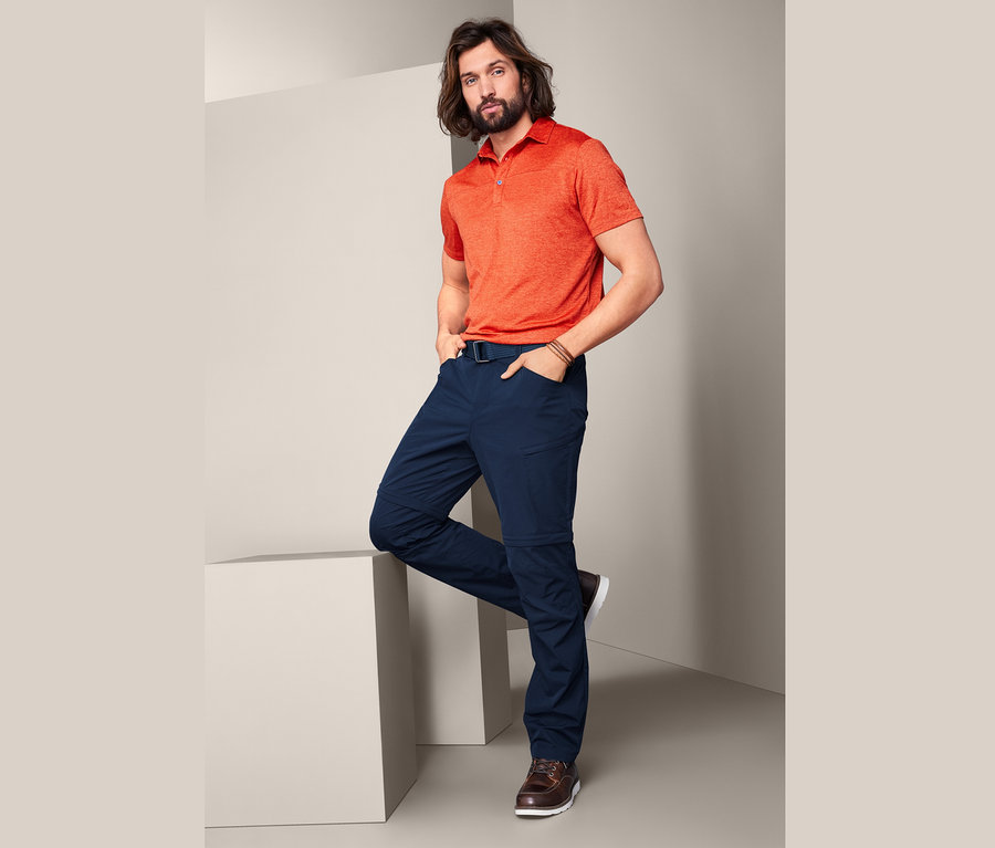 Mann posiert mit den Händen in den Taschen und einem Bein auf einem weißen Würfel. Er trägt ein orangefarbenes Poloshirt und eine dunkelblaue 2-in-1-Funktionshose.