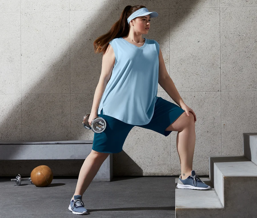 Frau macht Übungen mit einer Hantel, sie trägt blaue Long-Sweatshorts und 2 Tops.