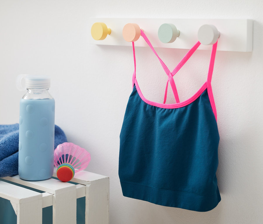 Ein Kinder-Sportbustier mit recyceltem Material hängt an einer Wandgarderobe. Daneben stehen eine Wasserflasche und ein Badmintonschläger.