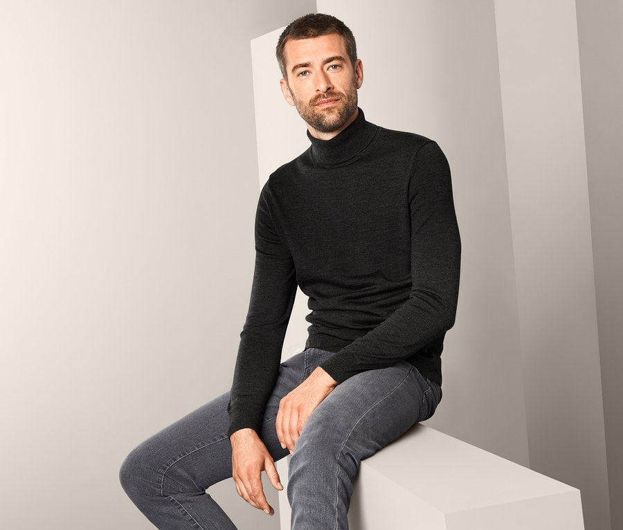 Mann sitzt und trägt einen anthrazitfarbenen Merino-Pullover.