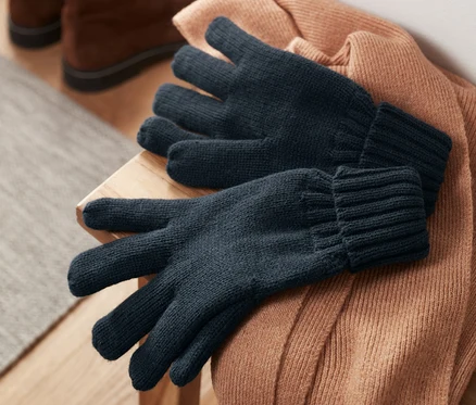 Strickhandschuhe liegen auf einem braunen Pullover, im Hintergrund Schuhe.