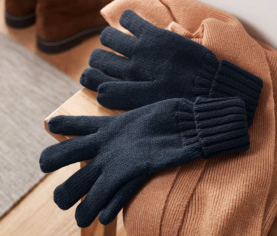 Strickhandschuhe liegen auf einem braunen Pullover, im Hintergrund Schuhe.