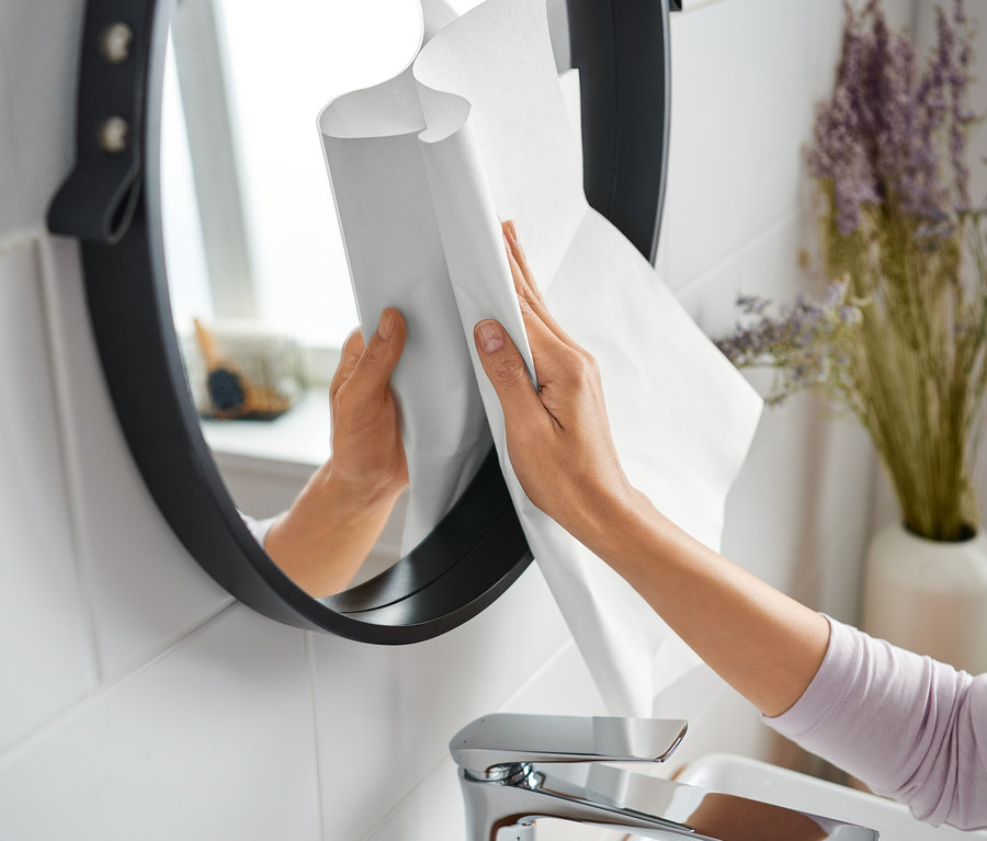 Eine Frau putzt mit einem Wundertuch einen runden Spiegel im Badezimmer.