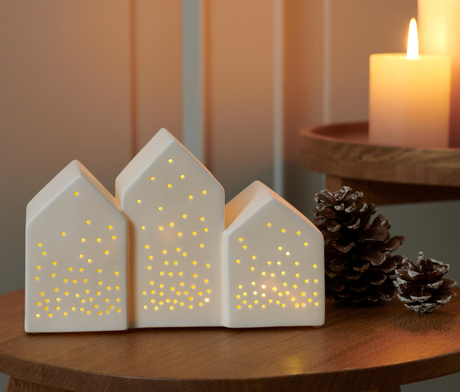 Ein LED-Dörfchen steht auf einem Holzhocker, daneben liegen Tannenzapfen. Im Hintergrund stehen zwei brennende Kerzen.