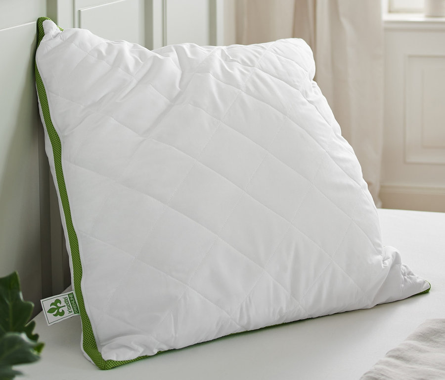 Ein weißes, gestepptes irisette® greenline Kopfkissen mit Klimaband, ca. 90 x 70 xm liegt auf einem Bett. Daneben liegt ein gefaltetes graues Handtuch.