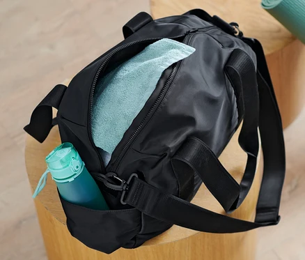 Eine schwarze Sporttasche liegt auf einem Holzhocker neben einer zusammengerollten Yogamatte.