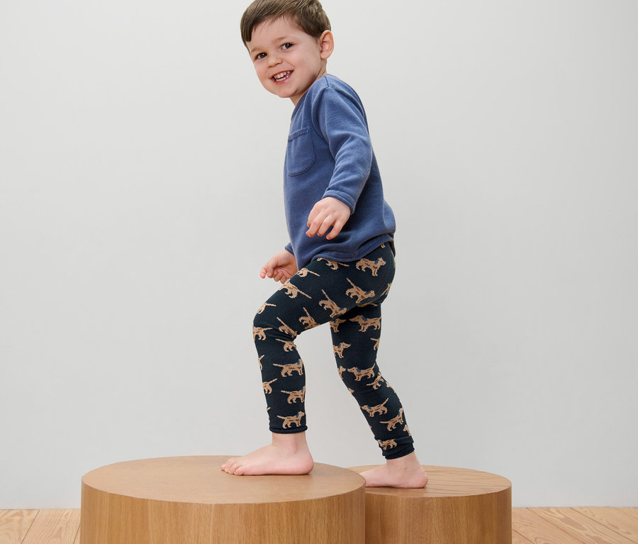Junge mit blauem Sweatshirt und 2 Kinder-Strickleggings mit Stickerei steht auf einem Holzpodest.