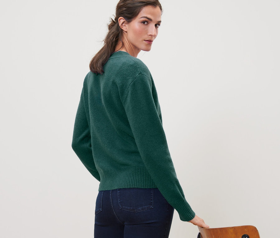 Frau blickt über die Schulter und trägt eine dunkelgrüne Strickjacke mit Wolle und Jeans.