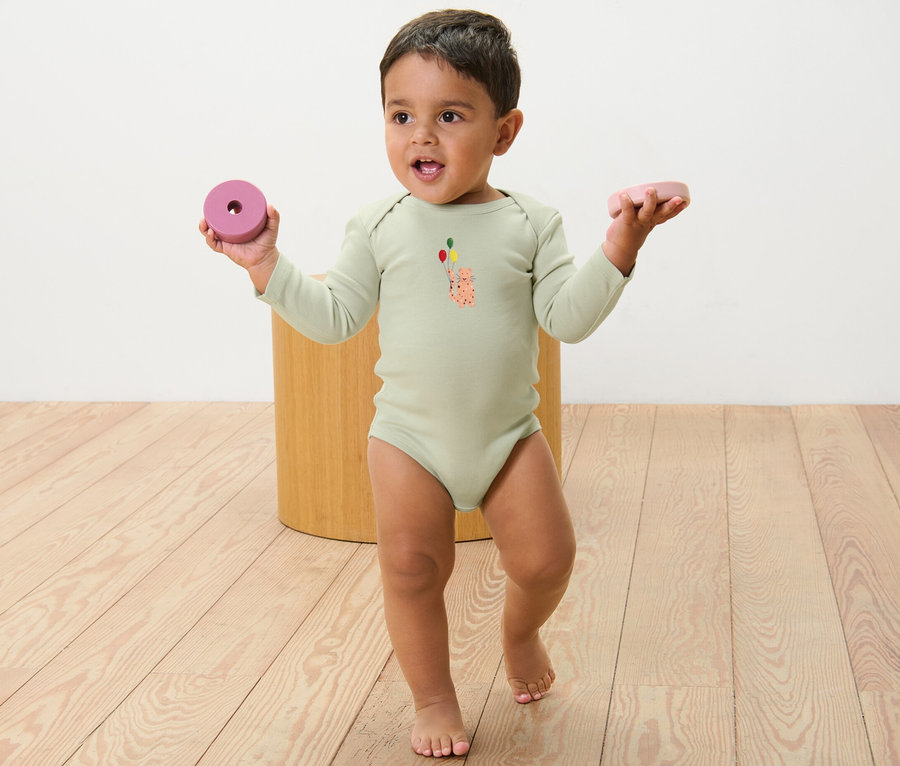 Kleinkind in einem langärmeligen Body mit Aufdruck, das in jeder Hand ein Spielzeug hält. 5 Baby-Bodys mit Mitwachsfunktion