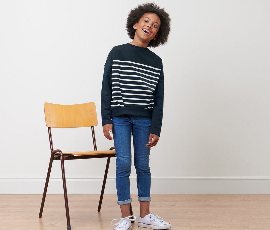 Ein lächelndes Kind steht neben einem Stuhl und trägt ein blaues Kinder-Sweatshirt.