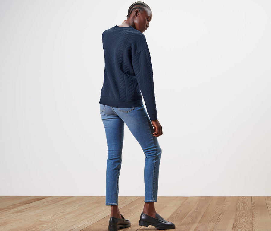 Frau posiert in blauem Sweatshirt mit Zopfmuster, Jeans und Schuhen.