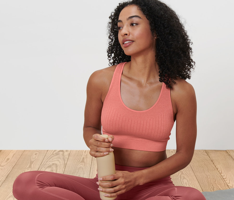 Eine Frau sitzt mit gekreuzten Beinen auf einer Yogamatte und hält eine Wasserflasche. Sie trägt ein altrosa Seamless-Sport-Bustier.