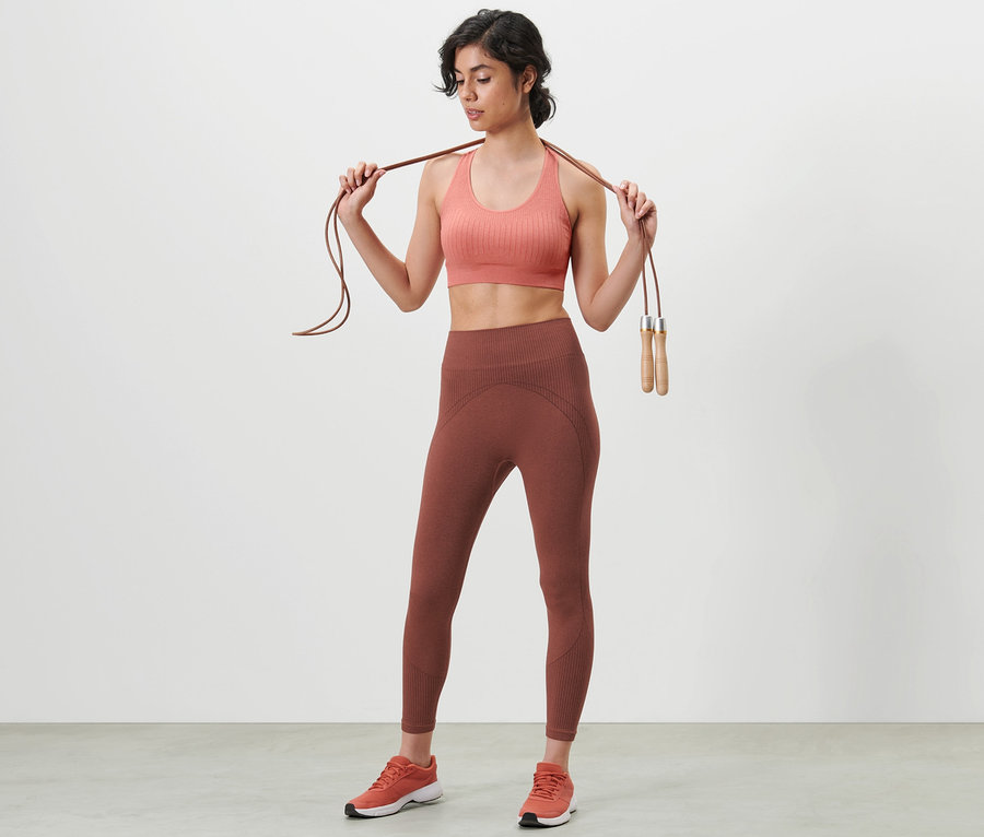 Frau hält ein Springseil auf ihren Schultern und trägt einen Sport-BH und eine 7/8-Seamless-Funktions-Sporttight.