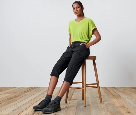 Frau sitzt auf einem Hocker und trägt eine Softshell-Funktionscapri und ein grünes T-Shirt.
