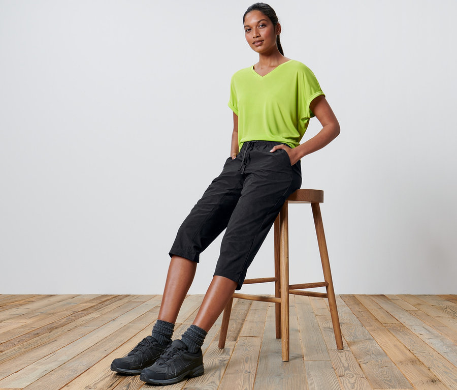 Frau sitzt auf einem Hocker und trägt eine Softshell-Funktionscapri und ein grünes T-Shirt.