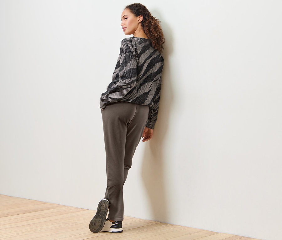 Frau lehnt an einer weissen Wand und trägt eine Joggpants und einen gemusterten Pullover.