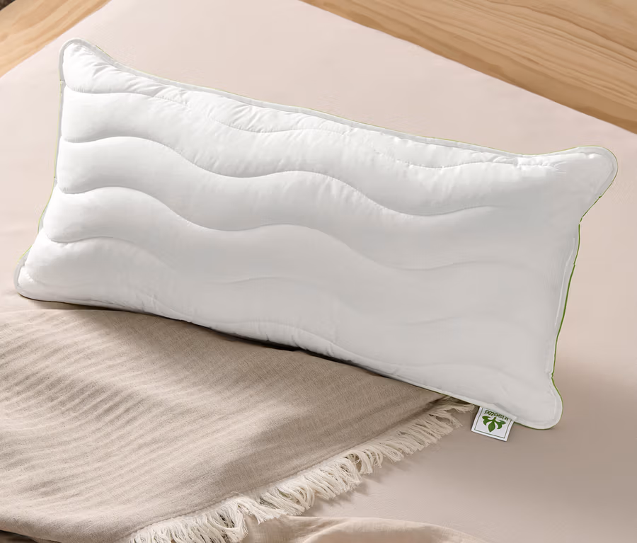 Ein weißes irisette® greenline Kopfkissen Megafil® BIO, ca. 40 x 80 cm liegt auf einer beigen Tagesdecke auf einem Bett.