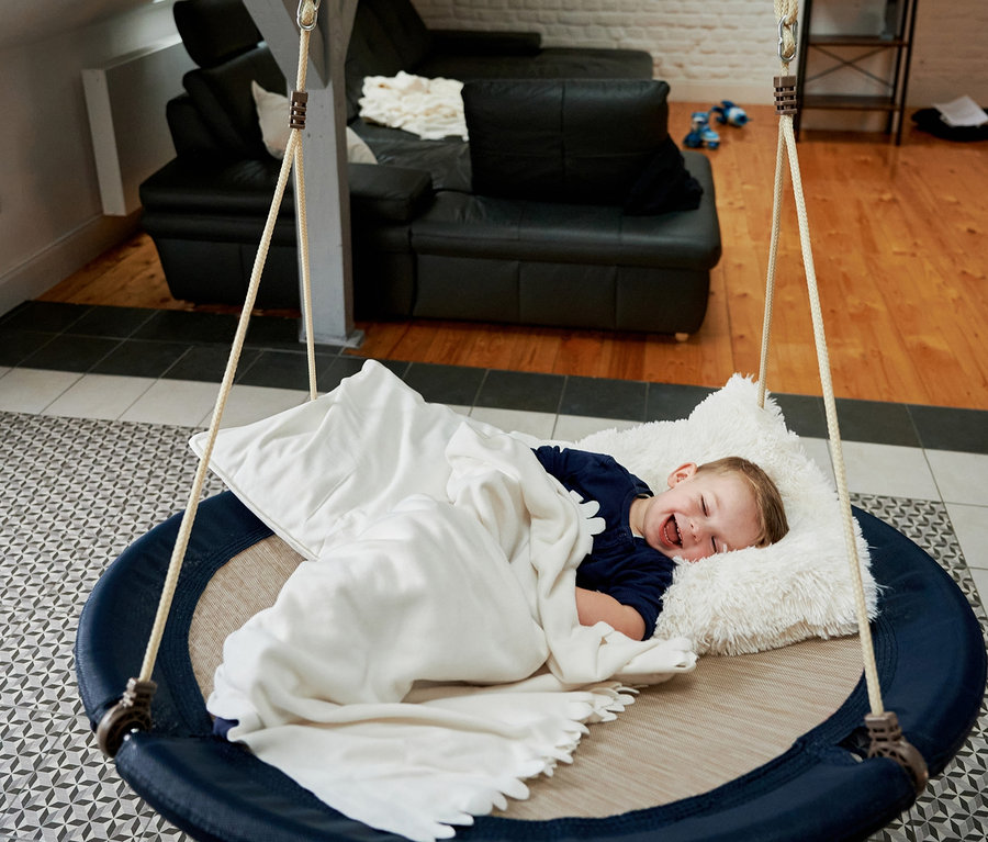 Ein Kind liegt auf einer HUDORA Nestschaukel »Lounge«, midnight, Ø 110 cm mit weißem Kissen und Decke.