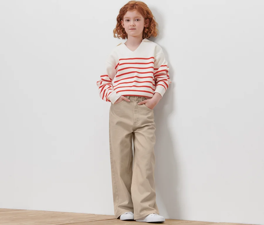 Mädchen steht an einer Wand in Kinder-Jeans – Fit »Lena«, einem gestreiften Pullover und weißen Turnschuhen.