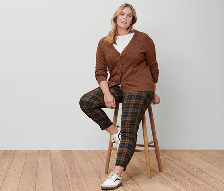 Eine Frau sitzt auf einem Hocker und trägt einen Feinstrickcardigan mit Zopfmuster, ein weißes Shirt mit 3/4-Arm, eine karierte Stretchhose und Leder-Sneaker.