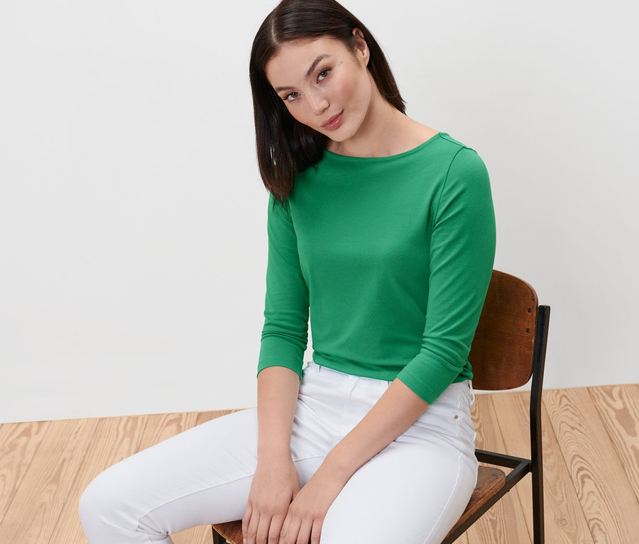 Frau sitzt auf einem Stuhl und trägt ein grünes Shirt mit 3/4-Arm.