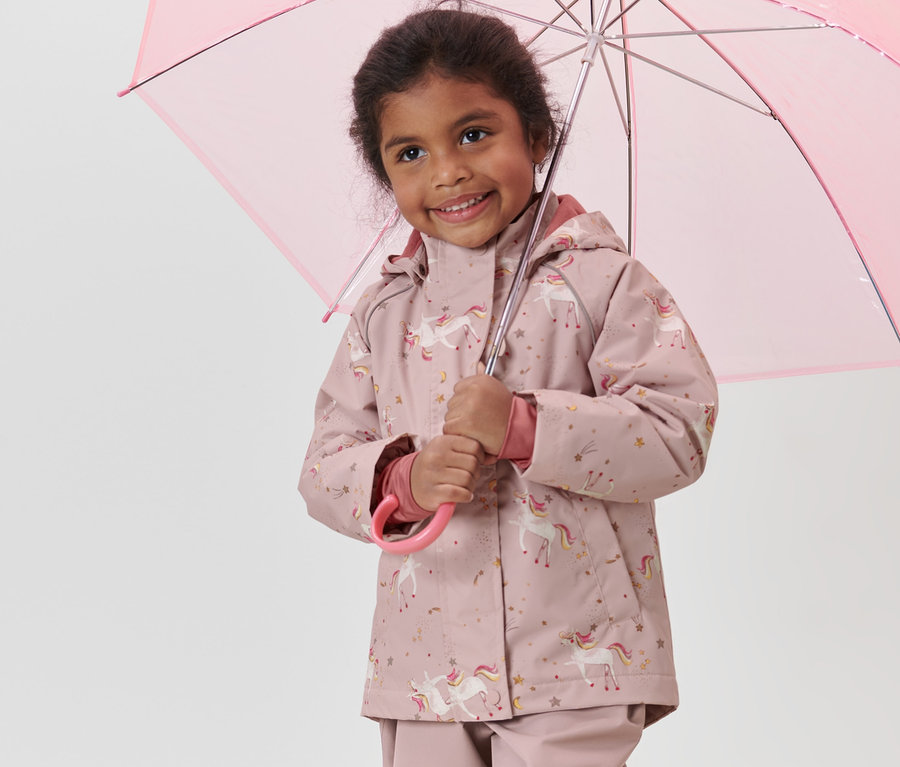 Lächelndes Mädchen hält einen rosa Regenschirm und trägt eine rosa Kinder-Thermo-Regenjacke mit Einhornmotiv.