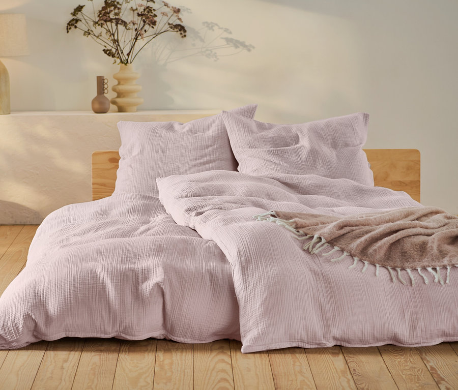 Rosafarbene Musselin-Bettwäsche in Normalgröße und Übergröße auf einem Bett mit Holzrahmen.