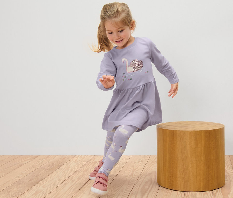 Lächelndes Mädchen in einem Kinder-Sweatkleid und Leggings mit Schwanenmuster springt auf einem Holzboden neben einem Holzhocker.