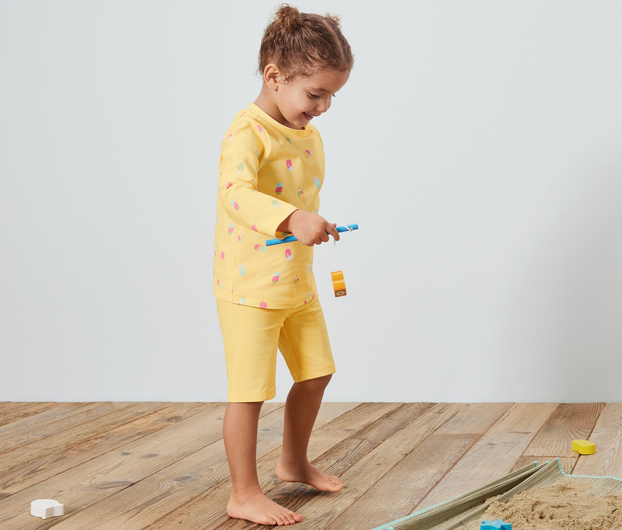 Mädchen spielt auf einer Decke mit Sand und gelbem Kinder-Kombi mit UV-Schutzfaktor 80.