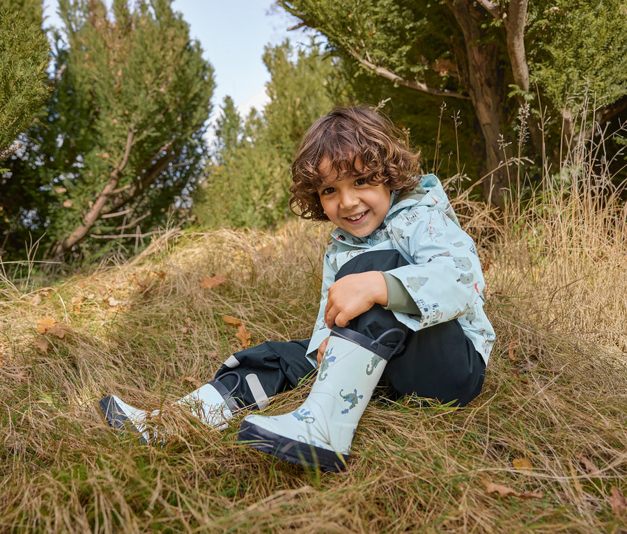 Ein Junge sitzt im Gras und trägt Kinder-Regenstiefel mit Drachen, eine Kinder-Regenjacke mit Rittern und eine Kinder-Regenhose.